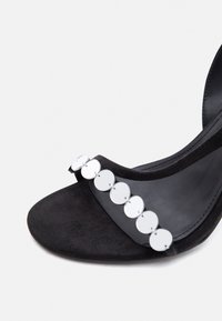 Μαύρο πέδιλο με ψηλό τακούνι και ανοιχτά δάχτυλα, με έναν μόνο λουρί που διαθέτει σειρά από κυκλικούς ασημένιους δίσκους.