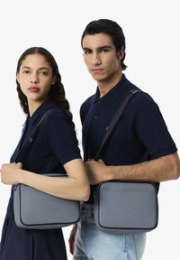 Borse a spalla Lacoste grigie, realizzate in materiale testurizzato, con logo in rilievo, dettagli laterali e tracolle regolabili.
