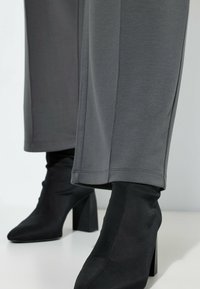 Czarne botki na wysokim obcasie o smukłym, dopasowanym fasonie, zestawione z szarymi, szerokimi spodniami o gładkiej fakturze i widocznych detalach przeszyć.