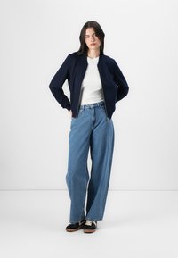 A young woman stands confidently with hands on her hips, wearing a navy jacket, white top, and wide-leg jeans, set against a plain background.