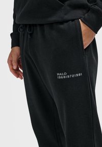 Schwarze Jogginghose aus weichem Material mit einem elastischen Bund, auf dem linken Oberschenkel steht der Text "HALO 1968|972|1981" in weiß.