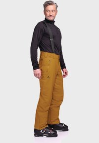 Men's ski pants in mustard yellow, featuring a loose fit, adjustable straps, multiple pockets, and black ski boots with red accents.