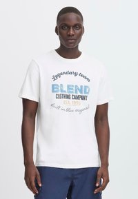 Balta medvilnės marškinėliai su mėlynu ir juodu tekstu. Dizainas apima "Blend Clothing Company" su retro stiliaus šriftu. Trumpomis rankovėmis, apvaliu kaklu.