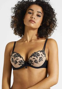 Soutien-gorge push-up noir et nude avec broderie florale noire, coques transparentes et bretelles réglables. Il présente des bords festonnés et un détail de nœud central.