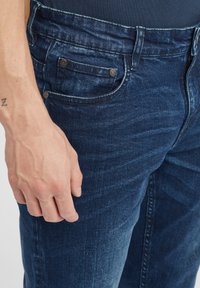 Temno modri kavbojasti hlače s izbledelim videzom, zasnova z petimi žepi, kovinske dodatke in šivane šive vzdolž noge.