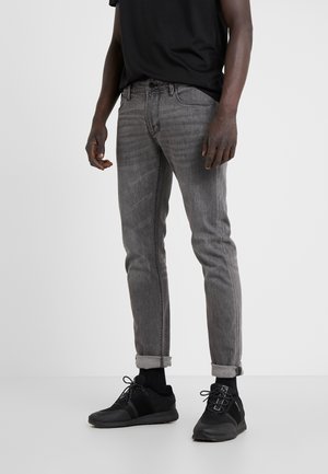 Mand iført grå slim-fit jeans, opsmøgede ved anklerne, sorte sneakers, sorte sokker og en sort t-shirt mod hvid baggrund.