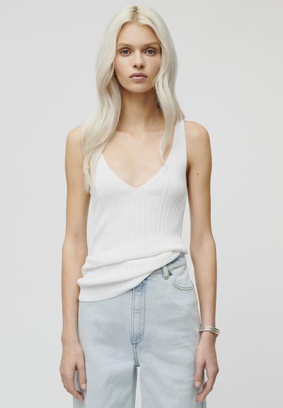 Fehér bordás camisole mély V-nyakú kivágással, világoskék farmernadrággal párosítva. Az ezüst karkötő a csuklón diszkrét kiegészítést ad.