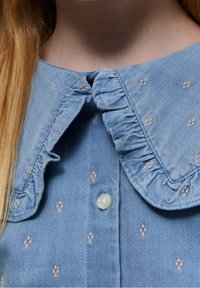 Blusa in denim azzurro chiaro con colletto arricciato, chiusura con bottoni madreperlacei e piccole, sottili fantasie floreali su tutta la stoffa.