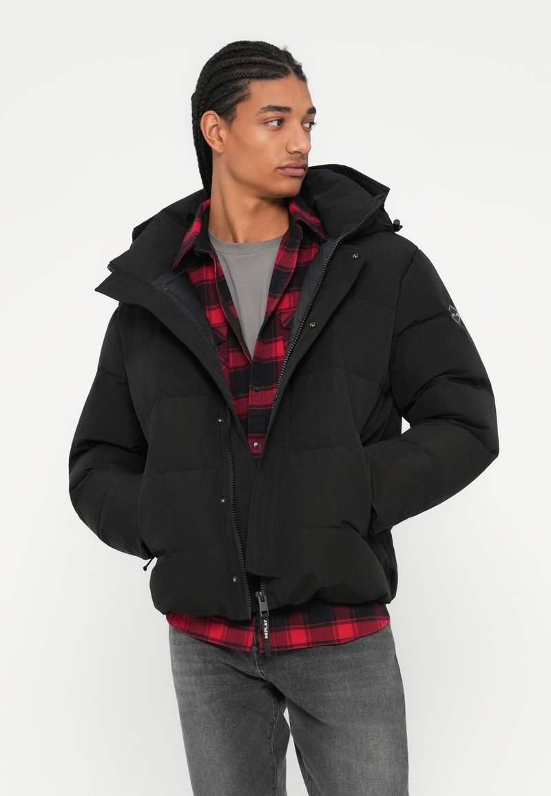 Schwarze Pufferjacke mit Kapuze, ausgestattet mit einem Reißverschluss und Seitentaschen, getragen über einem rot-schwarzen karierten Hemd, kombiniert mit grauen Jeans.