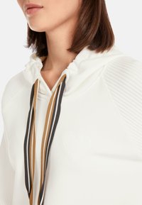 White hoodie with drawstrings in brown, black, and gold. Features ribbed texture on shoulders and an embossed heart design on the front.