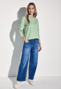 Ljusgrön våffelstickad tröja med långa ärmar, parat med vida blå denimjeans och vita sneakers, stående mot en vit bakgrund.