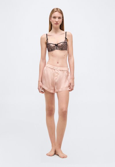 Νέα γυναίκα που στέκεται ξυπόλητη, φορώντας δαντελωτό μαύρο σουτιέν και ανοιχτό ροζ σατέν σορτς, πάνω σε απλό φωτεινό φόντο.