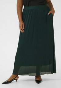 Falda maxi plisada de color verde oscuro con cintura elástica y bolsillos laterales, combinada con tacones de punta negra. Tejido ligero y texturizado.