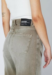 Jeans verde oliva de denim, con un bolsillo trasero y una etiqueta negra con texto blanco en la cinturilla. Detalles sutiles de textura y costura.