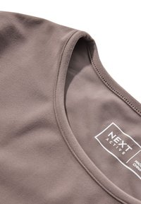 Top sportivo marrone con scollo rotondo, texture morbida, cuciture piatte e etichetta con logo, che evidenzia un design minimale e funzionale.