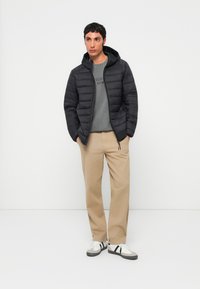 Schwarze Pufferjacke mit Kapuze, ausgestattet mit gestepptem Design und Reißverschlussdetails, über einem grauen Sweatshirt und beigen Hosen getragen, kombiniert mit Sneakers.