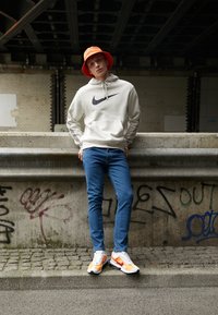 Moletom cinza da Nike com logo preto, jeans ajustados azuis e ténis branco-laranja. Chapéu de balde laranja brilhante. Cenário urbano com fundo de grafite.