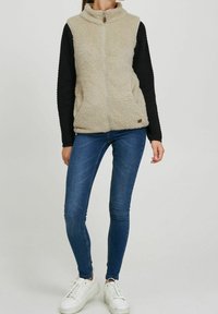 Beige Fleeceweste mit hohem Kragen und Reißverschluss, kombiniert mit schwarzen, strukturierten langen Ärmeln. Getragen mit blauen Skinny-Jeans und weißen Sneakers.