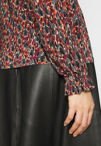 Bedruckte Bluse mit einer Mischung aus roten, orangenen und grauen Mustern, ausgestattet mit einem gerafften Ärmelabschluss, kombiniert mit einem schwarzen Lederrock.