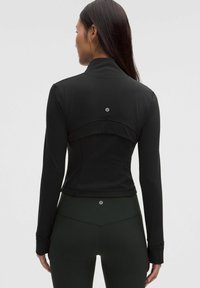 Crni atletski jakna s dugim rukavima, s visokim ovratnikom i dizajnom stražnjeg panela. Glatka tekstura, uski kroj, detalj logotipa na stražnjoj strani.