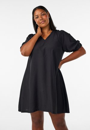 Frau mit glatten schwarzen Haaren, die ein kurzes schwarzes Kleid mit Puffärmeln trägt, steht mit einer Hand am Hals und der anderen an der Hüfte.