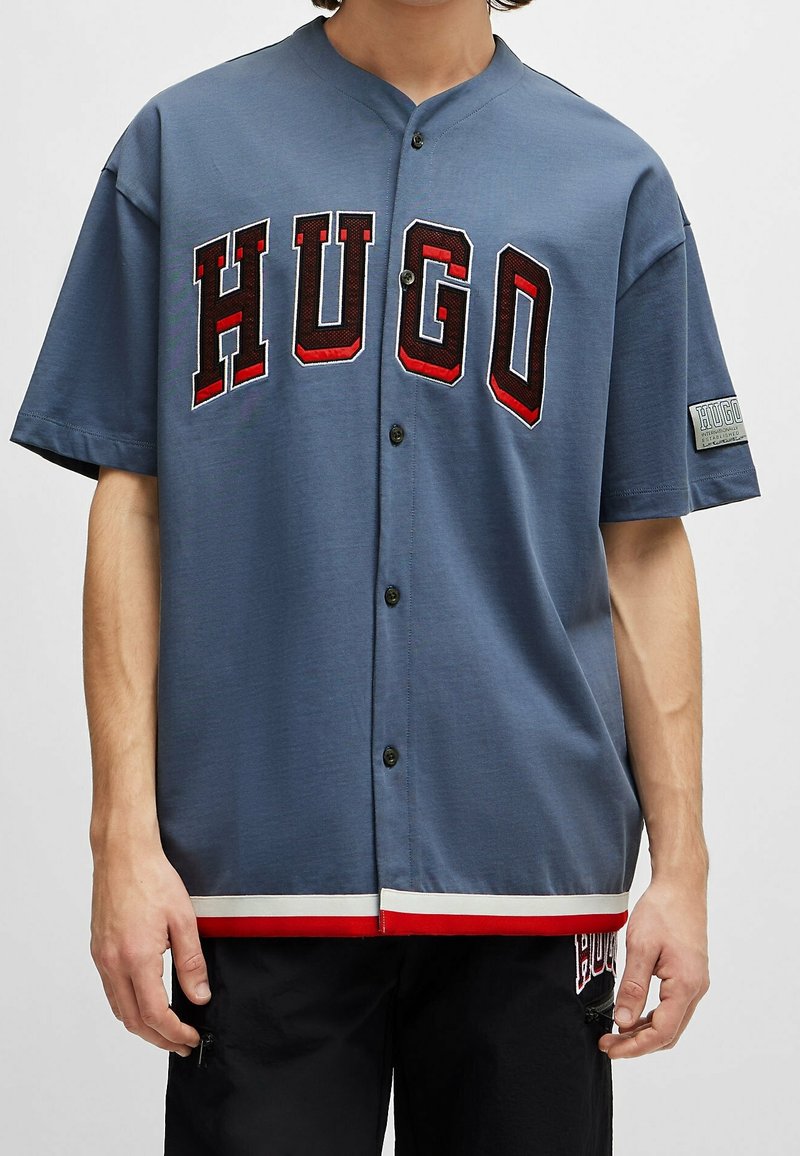 Personne portant une chemise bleue boutonnée avec de grandes lettres "HUGO" rouges et noires sur la poitrine et un écusson sur la manche gauche.