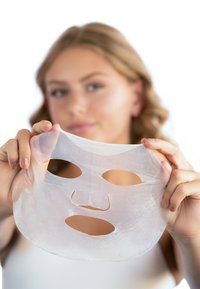 Doorzichtig gezichtsmasker met uitsparingen voor het gezicht, een gladde textuur en een glanzende afwerking, vastgehouden door een persoon tegen een vervaagde achtergrond.