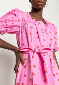 Robe rose avec des manches bouffantes courtes, une ligne de boutons et une ceinture nouée à la taille, ornée d'un motif de fraises. Tissu en coton doux.