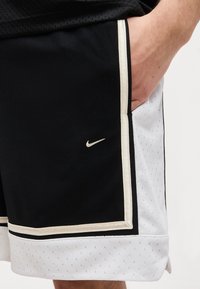 Gros plan sur la main d'une personne dans la poche d'un short de sport Nike noir et blanc avec une bordure beige et le logo Nike sur tissu noir.