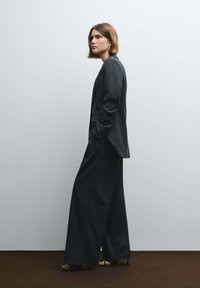 Costume sombre à fines rayures avec pantalon à jambes larges et blazer ajusté. Le tissu semble texturé. Le mannequin porte des sandales noires à talons hauts.