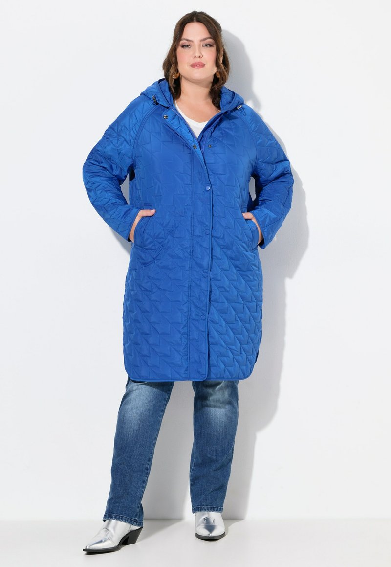 Blauer, gesteppter langer Mantel mit Kapuze, ausgestattet mit einer Knopfleiste, Seitentaschen und einem strukturierten Muster, getragen über blauen Jeans und silbernen Stiefeletten.