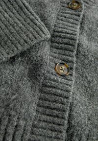 Harmaa villaneule, jossa on paksu ribbikuvio, käännettävä kaulus ja kontrastiväriset ruskeat napit. Pehmeä, pörröinen materiaali näkyvissä.