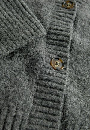 Hallhalli hallukka, millel on paks ribakujundus, allaklapitav kaelus ja kontrastsed pruunid nupud. Pehme, udune materjal on nähtav.