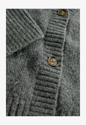 Hallhalli hallukka, millel on paks ribakujundus, allaklapitav kaelus ja kontrastsed pruunid nupud. Pehme, udune materjal on nähtav.