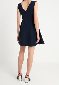 Námořnická modrá bezrukávová šaty s rozšířenou sukní, zadním výstřihem ve tvaru V a skrytým zipem. V kombinaci s černými lodičkami se stříbrným blokovým podpatkem.
