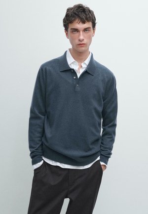 Dunkelgrauer Strickpullover mit langen Ärmeln und einem Kragen, der über einem weißen Hemd mit Kragen getragen wird und drei Knöpfe am Hals hat.