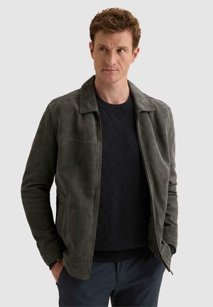 Homme aux cheveux courts et bouclés portant une veste en daim vert foncé par-dessus un pull en maille noir, regardant vers sa gauche avec les mains dans les poches.