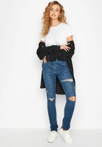 Camiseta blanca superpuesta bajo una camisa negra de gran tamaño, combinada con jeans skinny rasgados azules y zapatillas blancas.