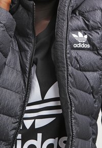 Svart puffjacka med ett kviltat mönster, dragkedja framtill och en vit Adidas-logotyp på bröstet, bärs över en svart tröja med en liknande logotyp.