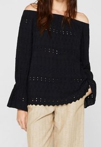 Sort off-the-shoulder bluse med broderet detaljer, lange klokkeærmer og en bølget kant. Båret med lyse stribede bukser.