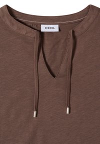 Chemise décontractée marron avec un col fendu et des cordons à embouts métalliques, comportant une étiquette cousue portant la mention "CECIL" à l'intérieur du col.