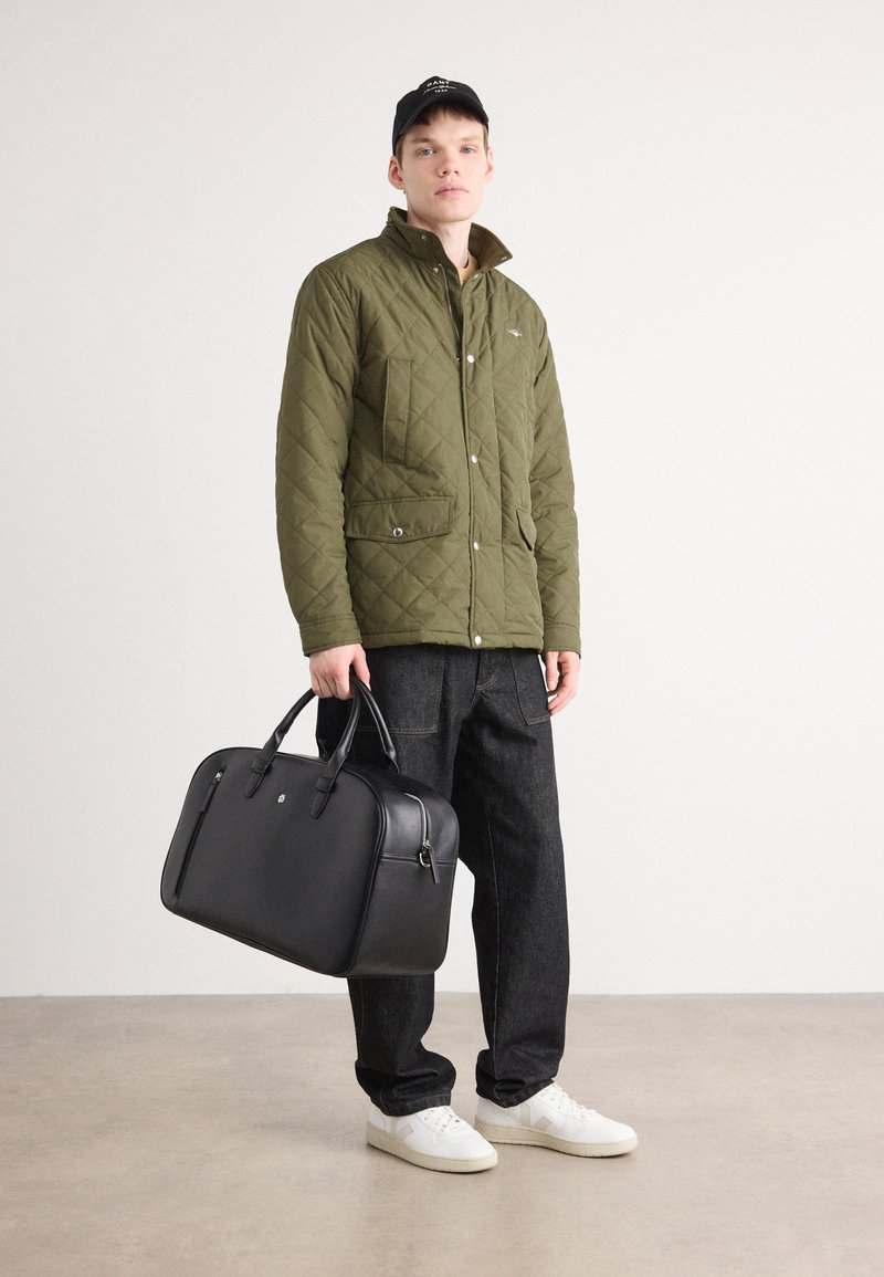 Czarna skórzana torba podróżna z podwójnymi uchwytami, pikowana oliwkowa kurtka zapinana na zatrzaski, ciemne dżinsy oraz białe trampki.