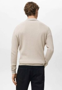 Beige geribbelde gebreide trui met lange mouwen en een ronde hals, gedragen over donkere broek. Zachte textuur, aansluitend ontwerp, minimale details.