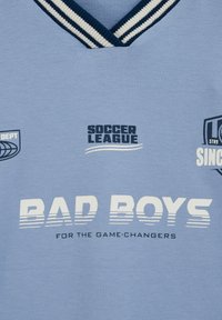 Vaaleansininen kangas, jossa tummansininen ja valkoinen raidallinen V-kaulus. Kauluksessa teksti: "SOCCER LEAGUE", "BAD BOYS" ja "FOR THE GAME-CHANGERS".