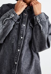 Persona che aggiusta il colletto di una camicia in denim nera oversize, lavata, con bottoni a scatto e tasche sul petto, su sfondo bianco.