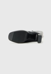 Bota de cuero negra con tacón cuadrado mostrada desde la suela sobre un fondo blanco liso.