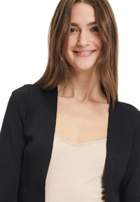 Fekete kardigán texturált kötésmintával, nyitott elülső résszel és hosszú ujjakkal, egy világos bézs ujjatlan felső fölé rétegezve. Sima szövésű anyag.
