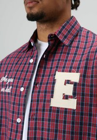 Rood en marineblauw geruit flanellen overhemd met witte knopen, voorzien van een geborduurde "E" patch en tekst met de woorden "See you in hell." Ontwerp met kraag.