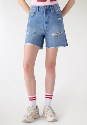 Persona che indossa pantaloncini di jeans blu usurati a vita alta, scarpe da ginnastica bianche e calzini bianchi con due strisce rosse, in piedi con le mani dietro la schiena.