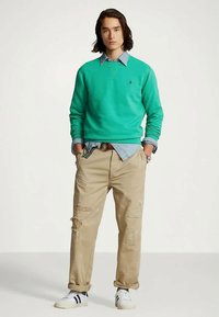 Νεαρός άνδρας με μεσαίου μήκους μαλλιά, φορώντας ένα πράσινο φούτερ, ανοιχτό μπλε πουκάμισο, μπεζ φθαρμένα παντελόνια και λευκά αθλητικά παπούτσια με μαύρες ρίγες.